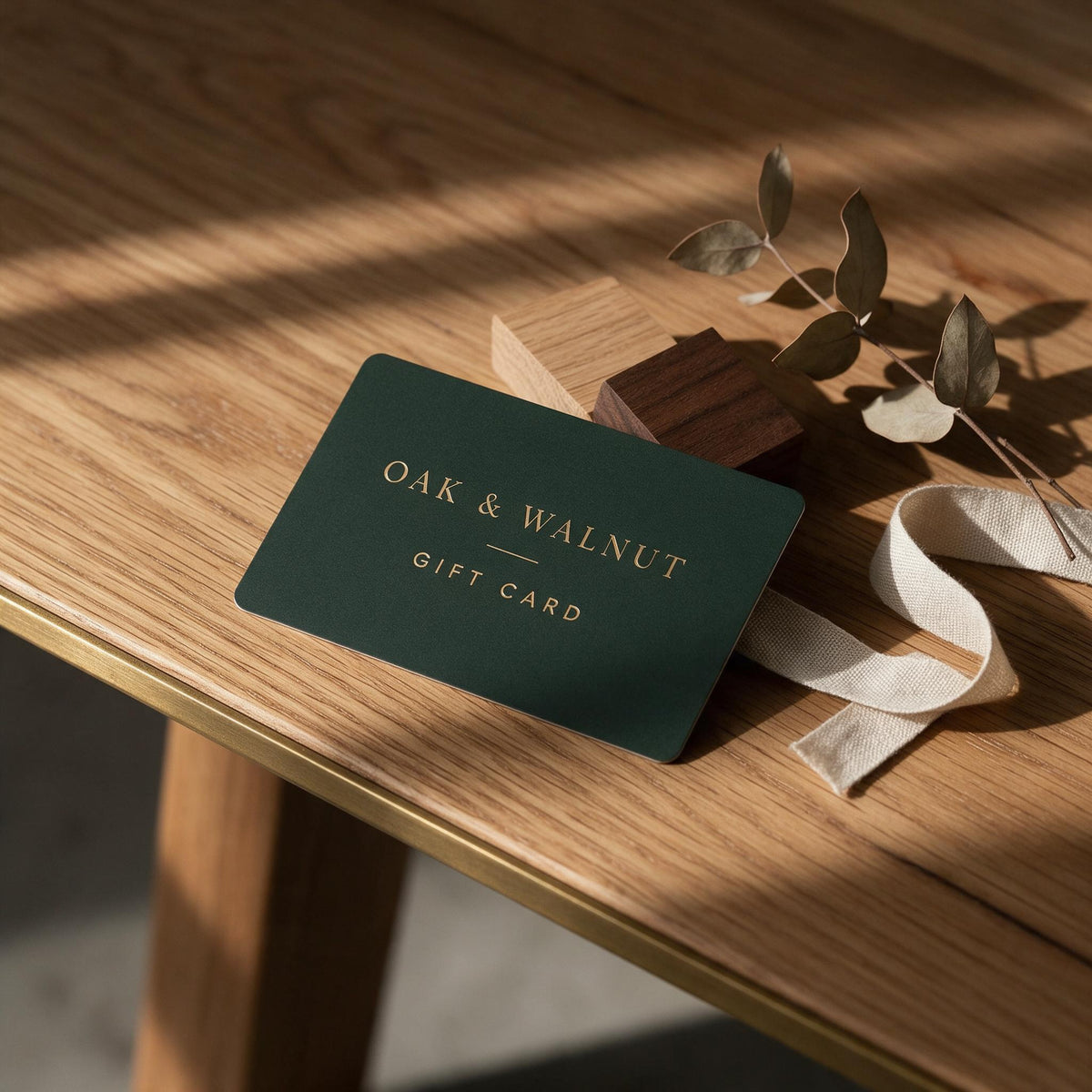 Oak & Walnut gift card in forest green with brass foil lettering, resting on a solid oak tabletop beside oak and walnut timber samples